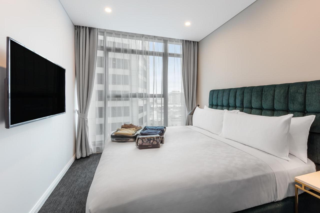 Meriton Suites Sussex Street, Sydney Kültér fotó The photo shows a modern hotel bedroom. The room features a large bed with a green upholstered headboard and crisp white linens. There are two pillows on the bed and a few folded items of clothing neatly placed on it. A flat-screen TV is mounted on t