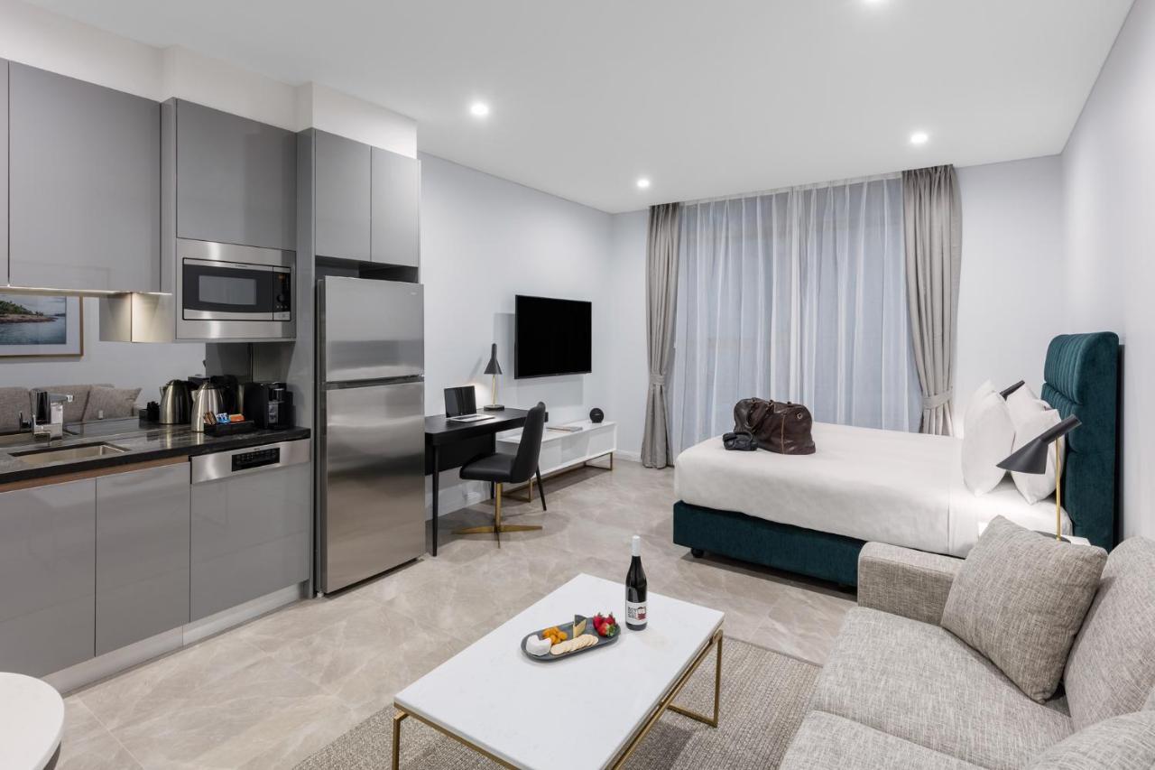 Meriton Suites Sussex Street, Sydney Kültér fotó The photo shows a modern studio apartment. On one side, there is a kitchenette with sleek gray cabinets, a refrigerator, and built-in appliances. In the living area, there is a comfortable gray sofa, a white coffee table with a fruit platter and a bo