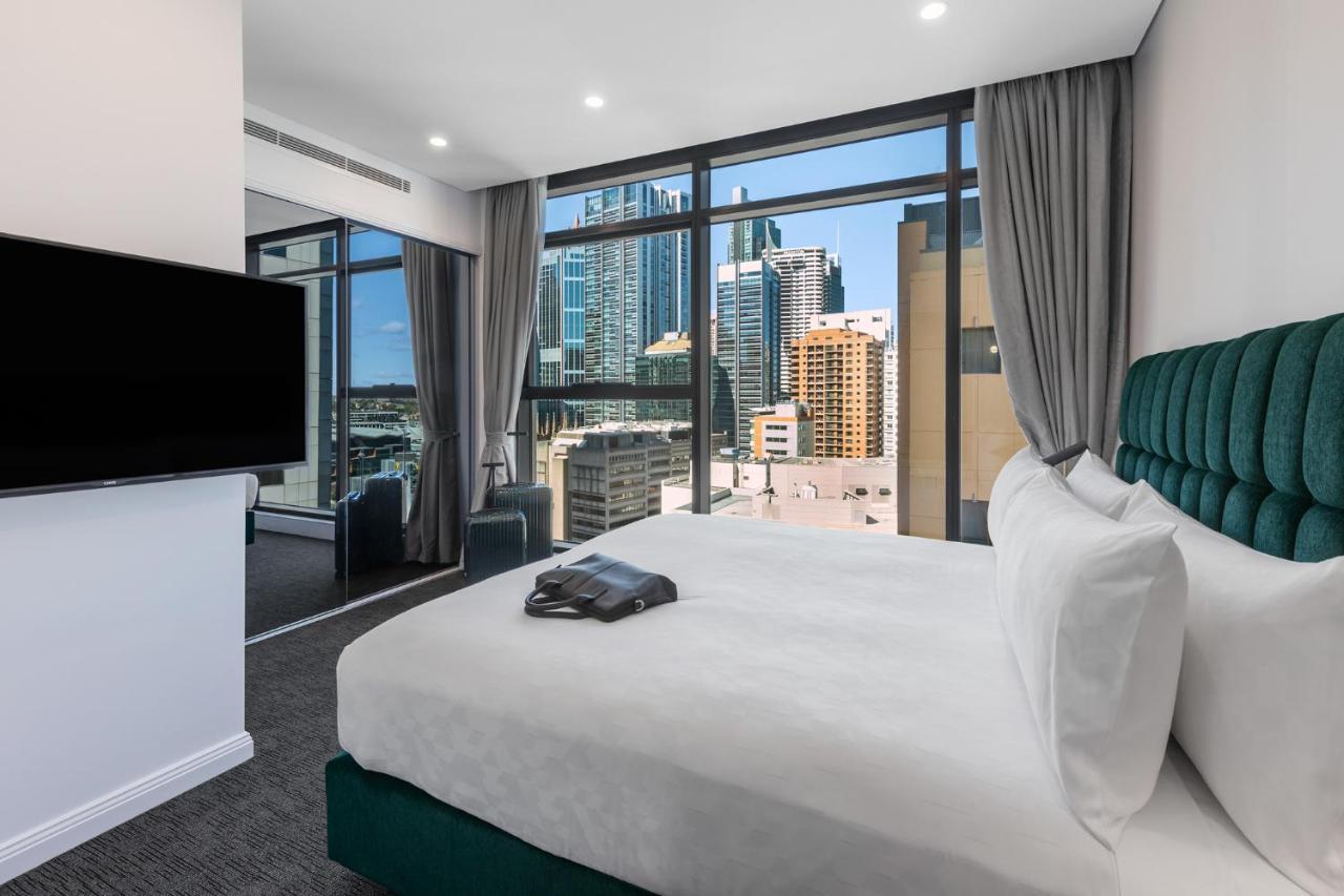 Meriton Suites Sussex Street, Sydney Kültér fotó The photo shows a modern hotel room featuring a large bed with white linens and a green upholstered headboard. There is a sleek television mounted on the wall opposite the bed, and the room has large windows that provide a view of a cityscape, includ