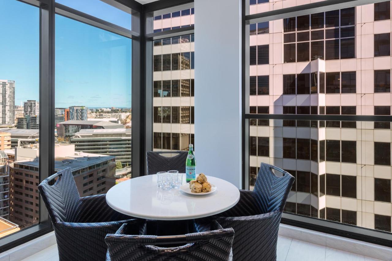 Meriton Suites Sussex Street, Sydney Kültér fotó The photo shows a modern dining space with a round white table set for four. There are four black woven chairs around the table. On the table, there's a bottle of sparkling water, two glasses, and a small plate with several cookies. The large windows