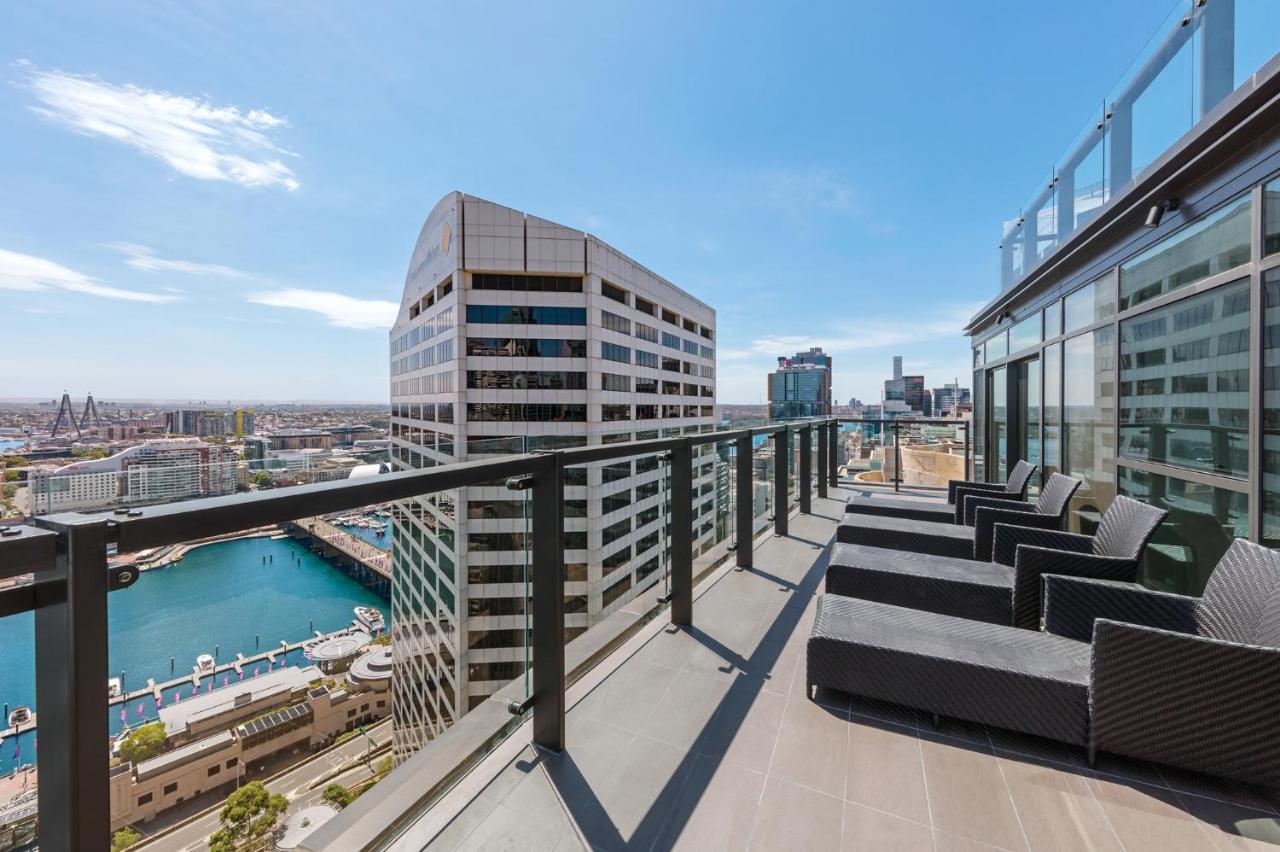 Meriton Suites Sussex Street, Sydney Kültér fotó The photo shows a balcony or terrace with a panoramic view of a city skyline. The balcony features modern outdoor furniture, including sleek lounge chairs. In the background, you can see a waterfront area with buildings and a harbor, along with a cle