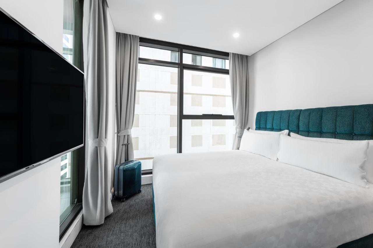 Meriton Suites Sussex Street, Sydney Kültér fotó The photo shows a modern hotel room featuring a large bed with a light-colored duvet and a blue upholstered headboard. There are two pillows on the bed. A large window allows natural light to enter the room, and gray curtains frame the window. Next t