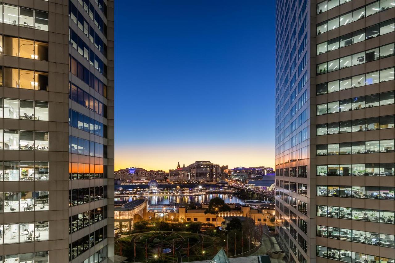 Meriton Suites Sussex Street, Sydney Kültér fotó The photo shows a view between two tall buildings, likely office towers, during twilight. On one side, there are glass windows reflecting the evening sky that transitions from blue to orange near the horizon. In the background, a cityscape is visible