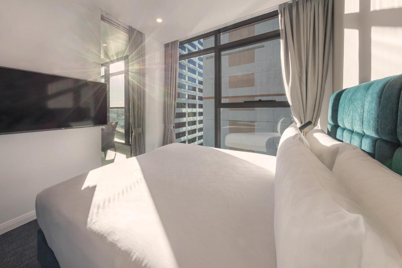 Meriton Suites Sussex Street, Sydney Kültér fotó The photo shows a modern hotel room with a neatly made bed featuring white bedding and a plush, tufted headboard in a shade of blue. Natural light is streaming through large windows, which are covered partially by light gray curtains. A flat-screen t