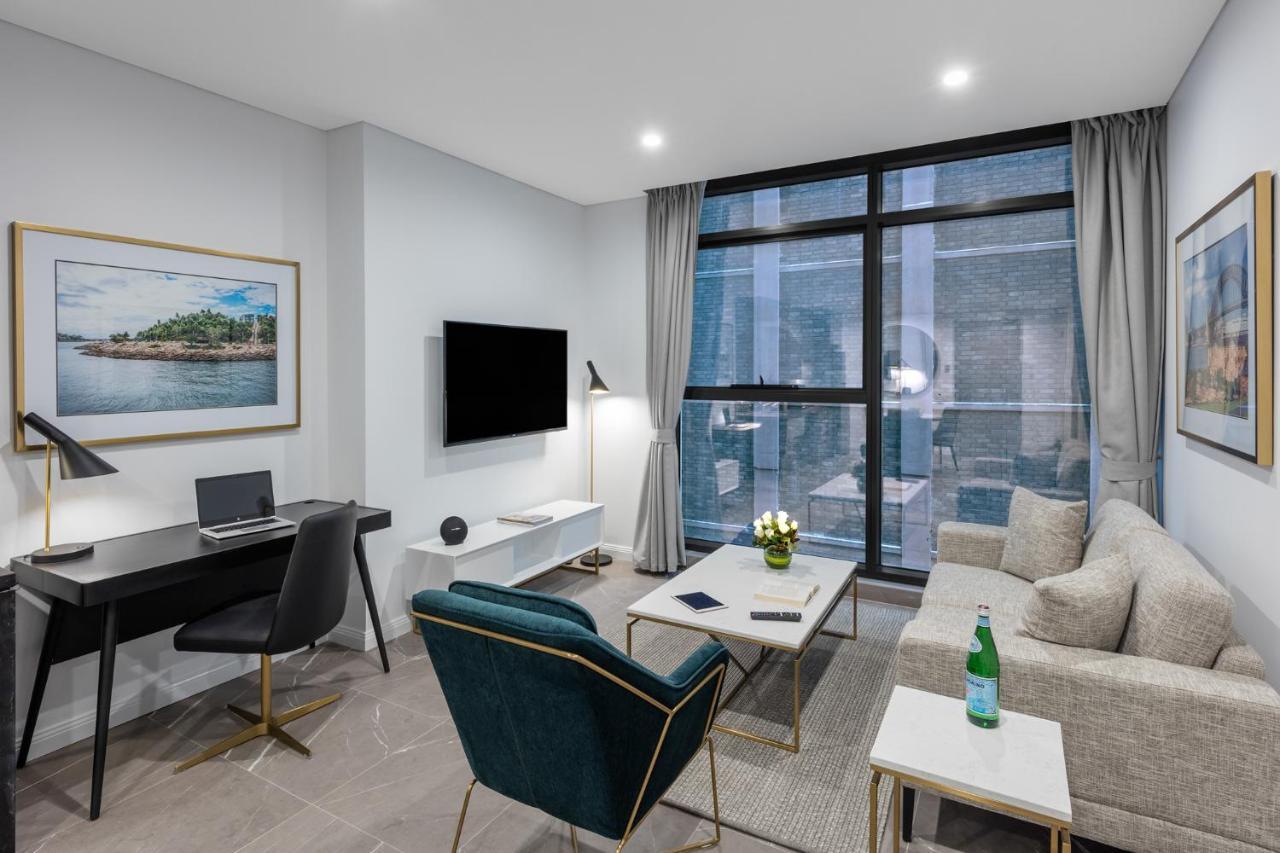Meriton Suites Sussex Street, Sydney Kültér fotó The photo shows a modern living area with a light color scheme. There is a comfortable seating arrangement that includes a beige sofa and an accent chair in teal. A coffee table is positioned in front of the sofa, with a small vase of flowers on it. 