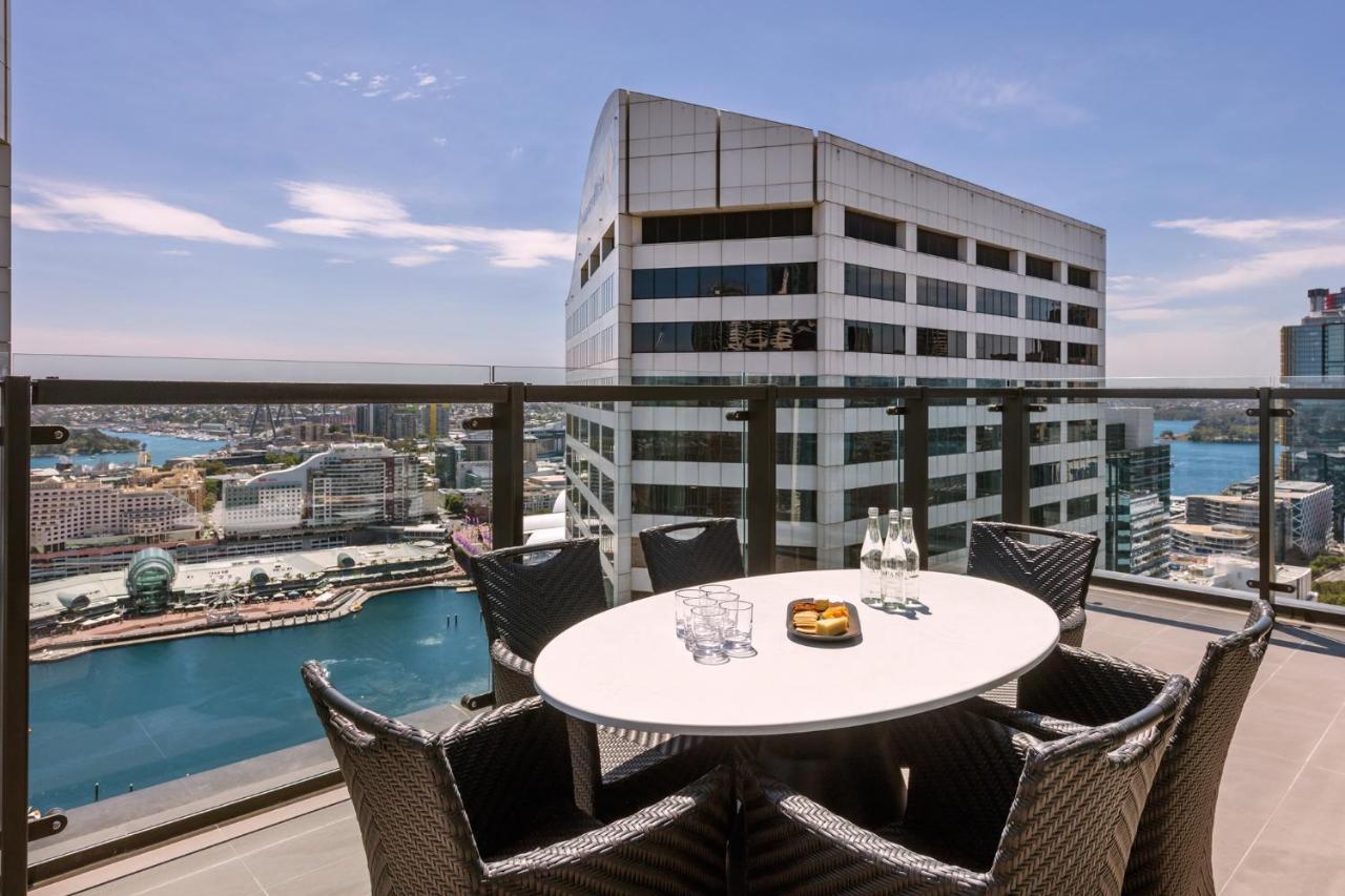 Meriton Suites Sussex Street, Sydney Kültér fotó The photo shows a balcony or terrace with a round table and several chairs arranged around it. The table is set with some drinks and a plate, possibly containing food. In the background, there is a view of a waterway and several buildings, including 