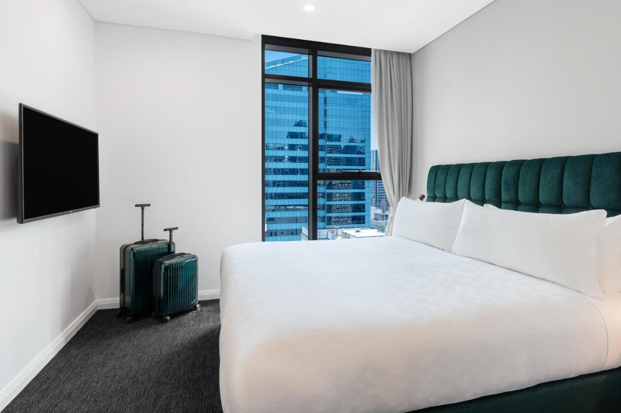 Meriton Suites Sussex Street, Sydney Kültér fotó The photo shows a modern hotel room featuring a large bed with white bedding and a green upholstered headboard. There are two green suitcases placed beside the bed. A flat-screen TV is mounted on the wall opposite the bed. The room has a large window