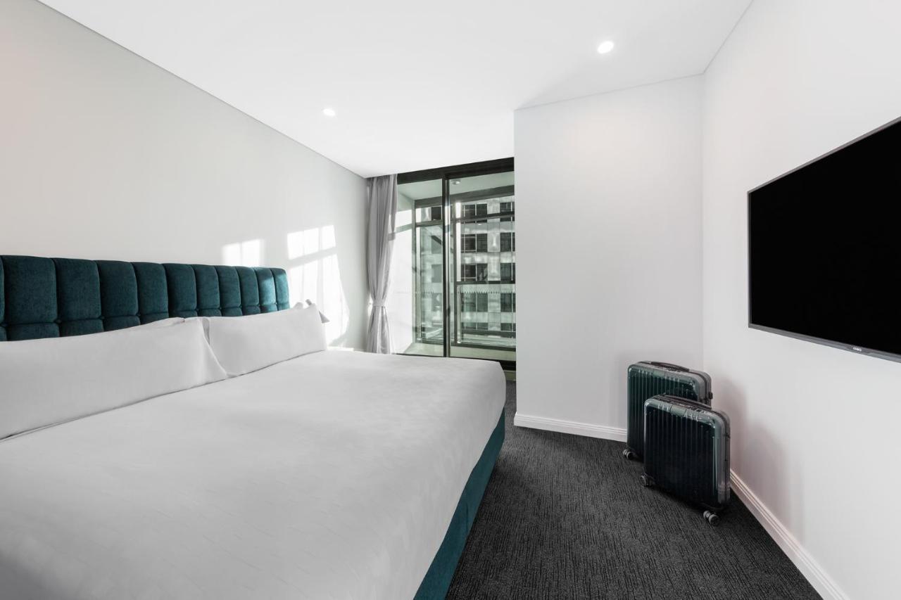 Meriton Suites Sussex Street, Sydney Kültér fotó The photo shows a modern hotel room featuring a large bed with a headboard in a teal color. The bedding is white and appears neatly arranged. There is a large window allowing natural light to enter, offering a view of an outdoor area. A flat-screen T
