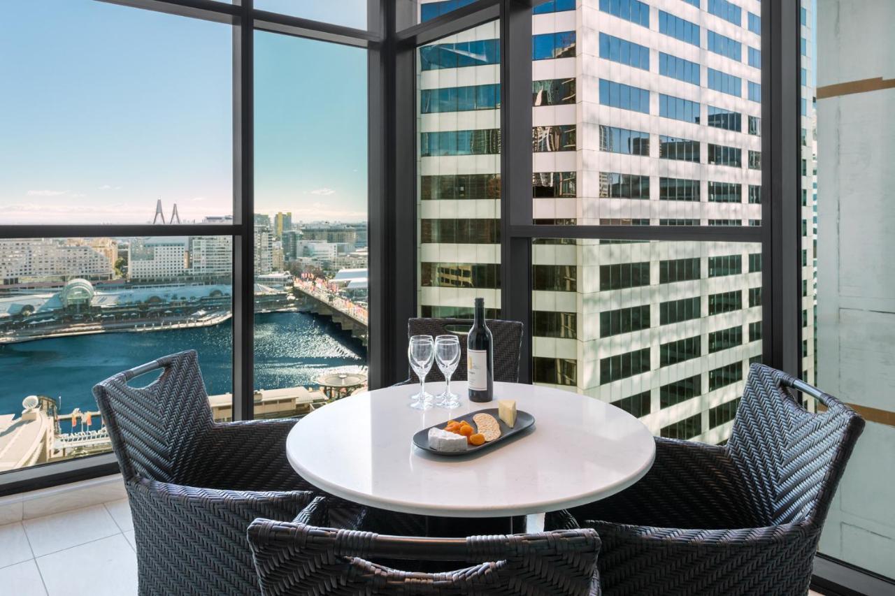 Meriton Suites Sussex Street, Sydney Kültér fotó The photo shows a modern dining area with a round table and four woven chairs. On the table, there are two wine glasses, a bottle of wine, and a plate with food items. The large windows provide a view of a cityscape featuring tall buildings and a bod