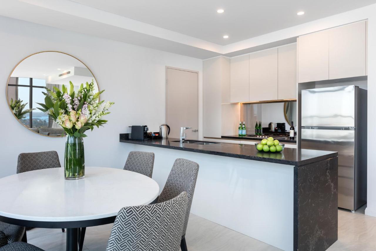 Meriton Suites Sussex Street, Sydney Kültér fotó The photo shows a modern kitchen and dining area. In the foreground, there's a circular dining table surrounded by four stylish chairs. A large vase filled with fresh flowers is placed on the table, adding a touch of color and elegance. 

In the kitc