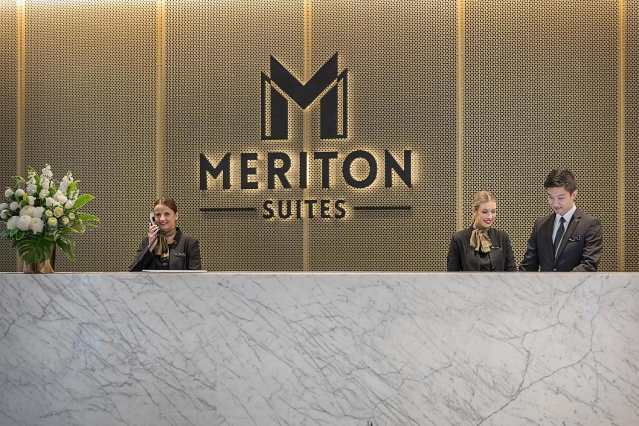 Meriton Suites Sussex Street, Sydney Kültér fotó The photo depicts the reception area of a hotel called "Meriton Suites." In the background, there is a large, illuminated sign featuring the hotel’s logo. The reception desk is made of marble and is staffed by two individuals dressed in professional 