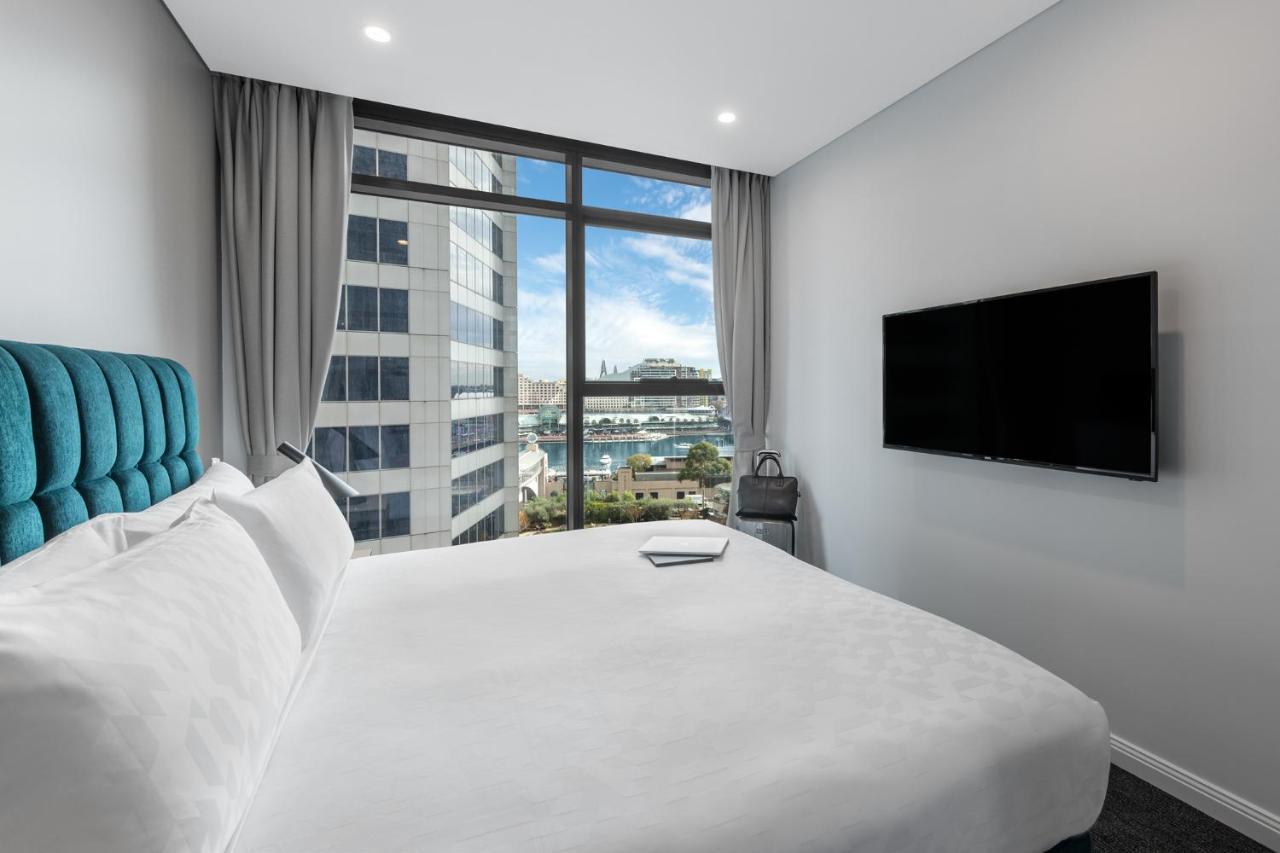 Meriton Suites Sussex Street, Sydney Kültér fotó The photo shows a modern hotel room with a large bed featuring a white comforter and pillows. There is a blue headboard that adds a pop of color to the decor. A large window provides a view of the outside, which includes buildings and a glimpse of wa