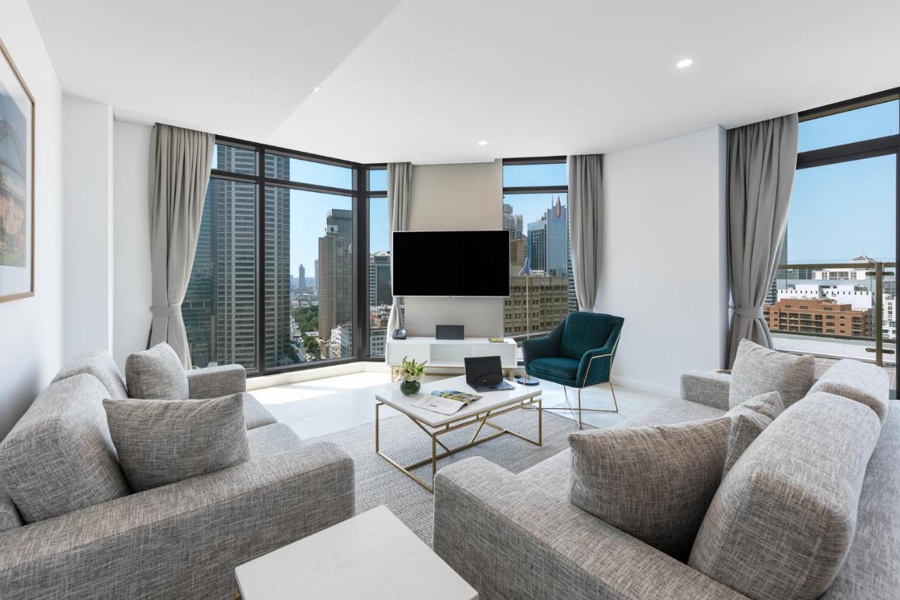 Meriton Suites Sussex Street, Sydney Kültér fotó The photo showcases a modern living room with large windows offering a view of a city skyline. The room features a light color palette with gray sofas arranged around a coffee table. A flat-screen TV is mounted on the wall, and there is a green armch